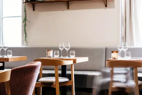 Stylishly set tables in a modern restaurant with wooden chairs, a padded bench, and elegant table decor featuring wine glasses and candles. Natural light streams through the window, creating a warm and inviting ambiance.